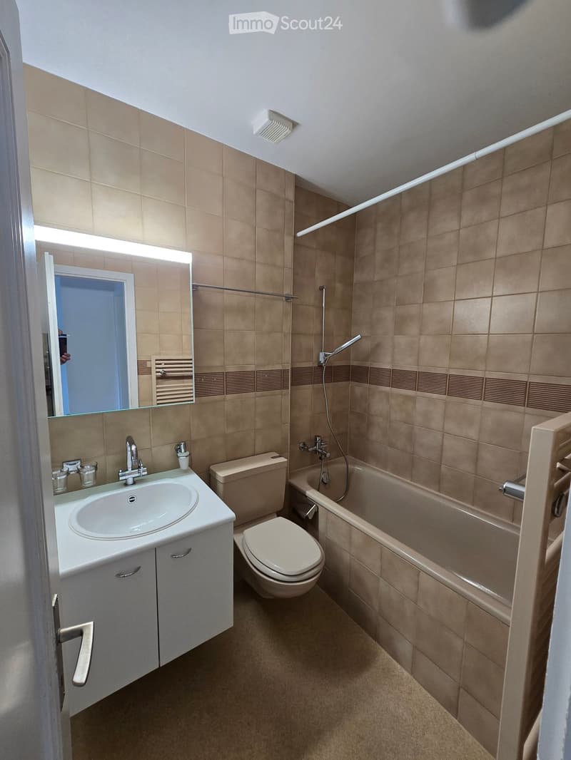 Modern bathroom with beige tiles, a white sink with a mirror, a toilet, a bathtub, and a towel rack.