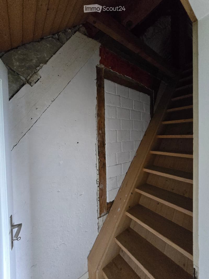 Wooden staircase leading up, white walls, exposed brick section, unfinished ceiling with visible beams and insulation.