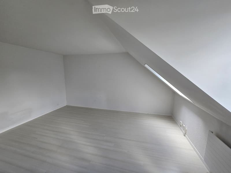 Empty attic space with wooden flooring, white walls, and a slanted ceiling with built-in lighting.