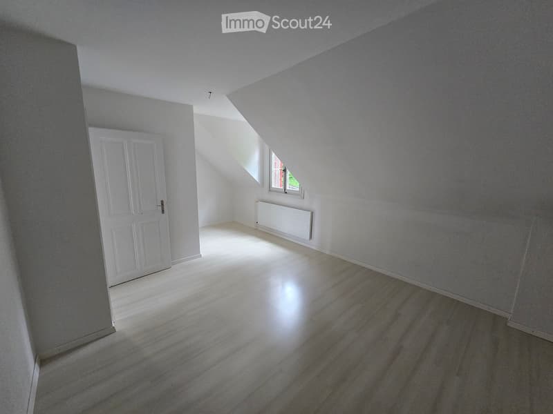 Empty attic room with wooden flooring, white walls, a white door, and a small window letting in natural light.