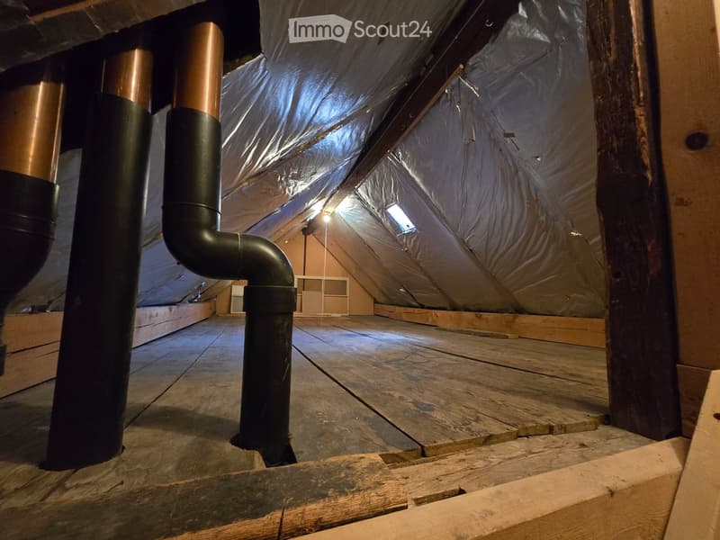 An attic space with wooden flooring, exposed wooden beams, insulation, and a black pipe running through it. The area is unfinished and appears to be under construction or renovation.