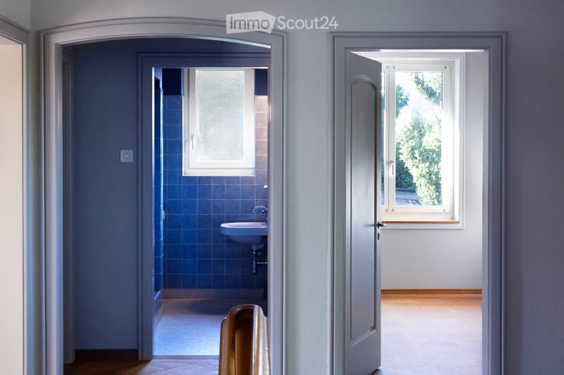 A hallway with light grey walls and wooden flooring, leading to a bathroom with blue tiles and a sink, and a room with a window showing greenery outside.