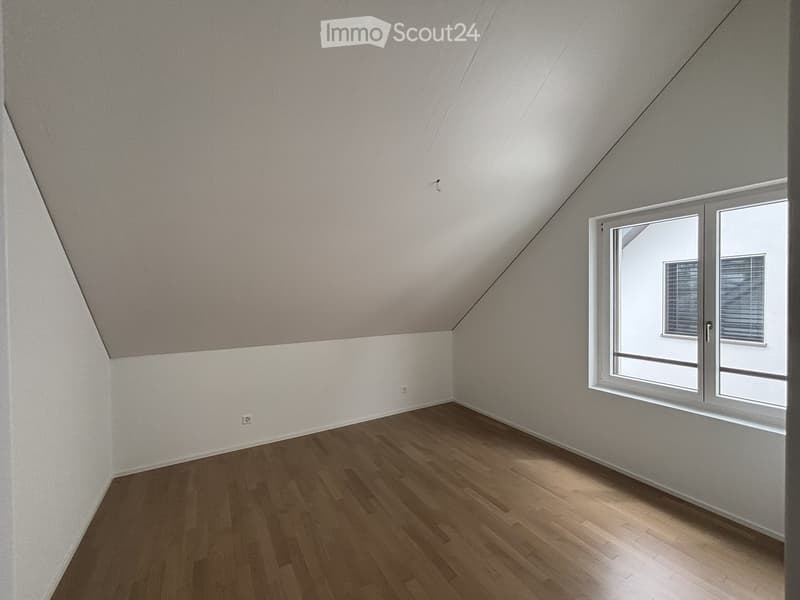 Empty attic room with wooden flooring, white walls, and a large window with blinds.