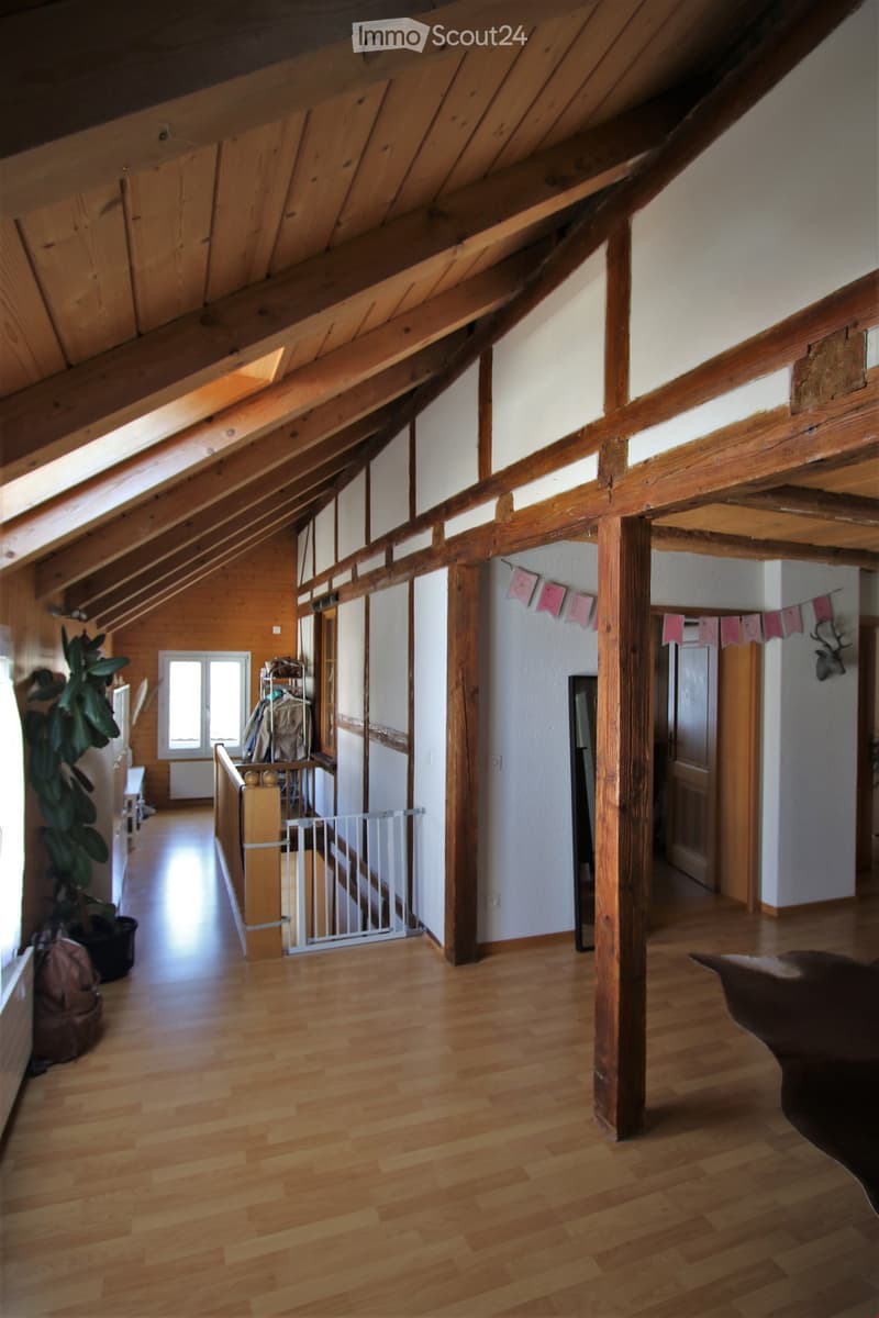 Spacious hallway with wooden beams and polished wooden flooring, leading to various rooms. Features include a staircase with a safety gate, a large potted plant, and a decorative deer head on the wall.