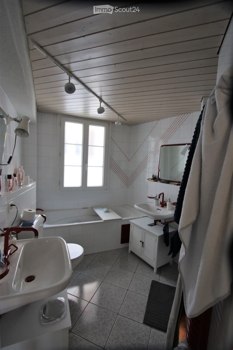 Bathroom with white tiled walls and floor, featuring a bathtub, two sinks, a toilet, a mirror, and a towel hanging on the side. The room has a slanted wooden ceiling with built-in lighting and a window providing natural light.