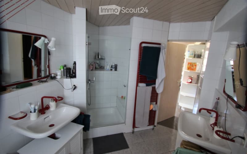 The image shows a small, well-lit bathroom with white tiled walls and a slanted wooden ceiling. It features a double sink setup with red faucets, a large mirror above one sink, and a glass-enclosed shower with a red towel rack. The bathroom includes a tow
