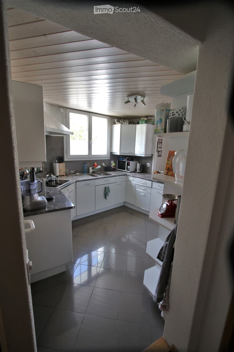 Modern kitchen with white cabinets, tiled floor, and various appliances including a sink, microwave, and blender. Natural light streams in from windows.