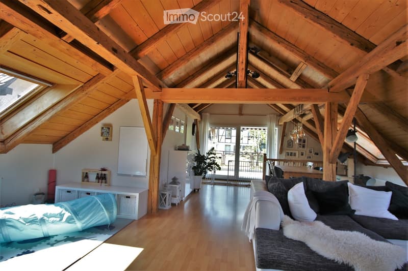 Bright, open living space with wooden beams and exposed ceiling, featuring a grey sectional sofa, coffee table, dining area, and large windows with a view of the balcony.