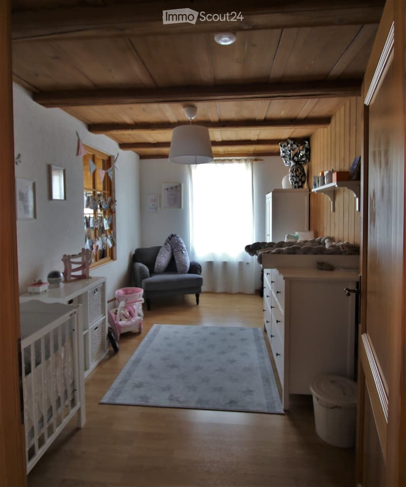 A cozy living room with wooden floors, a grey sofa, a white cradle, a cabinet, a vase on top, a small table, and a rug.