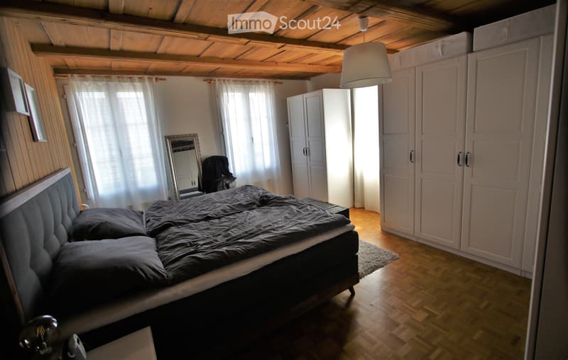 Cozy bedroom with wooden floors, a large bed with dark bedding, white wardrobes, a hanging lamp, and a mirror on a stand.