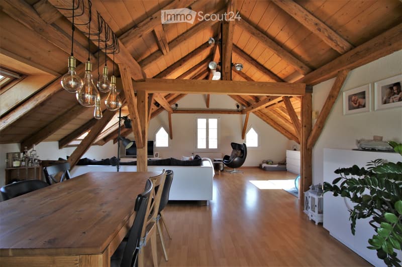 Open-plan living and dining area with exposed wooden beams, large windows, and modern furniture including a dining table, chairs, sofa, and a TV screen.
