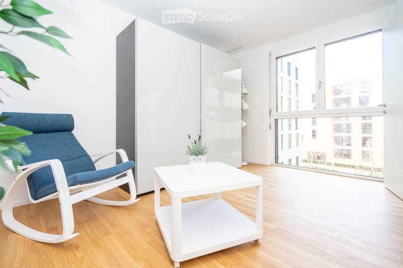 Modern living room with a white rocking chair, white coffee table, potted plant, and large white wardrobe. Large windows offer a view of the city.