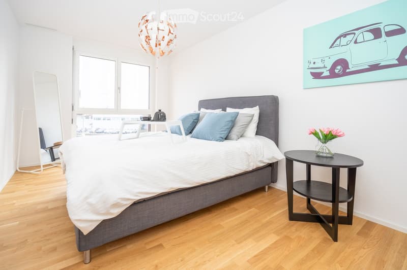 Modern bedroom with a grey upholstered bed, white bedding, blue pillows, a nightstand with a vase of pink flowers, a mirror, and a stylish pendant light fixture.