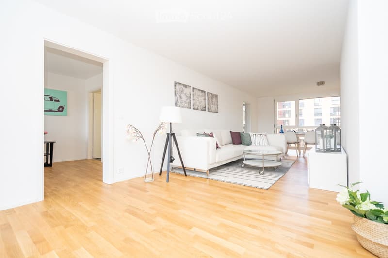 Bright living room with white walls, wooden flooring, white sectional sofa, coffee table, floor lamp, and decorative vases.