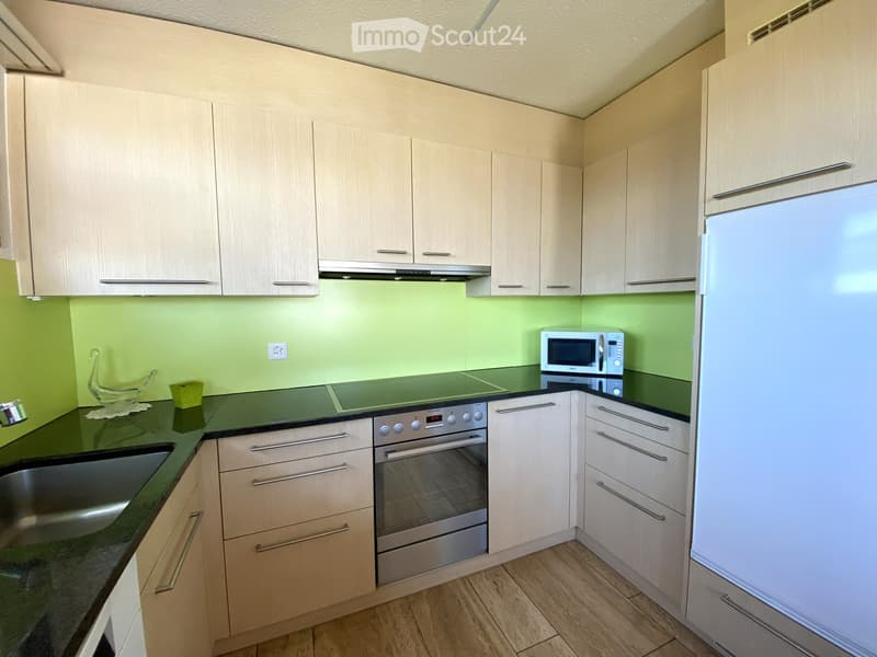 Modern kitchen with light wood cabinets, stainless steel appliances, and a green backsplash.