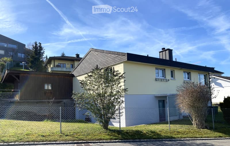 Two-story house with a combination of beige and dark brown exterior walls, multiple windows, a chimney, and a fenced front yard with trees and shrubs.
