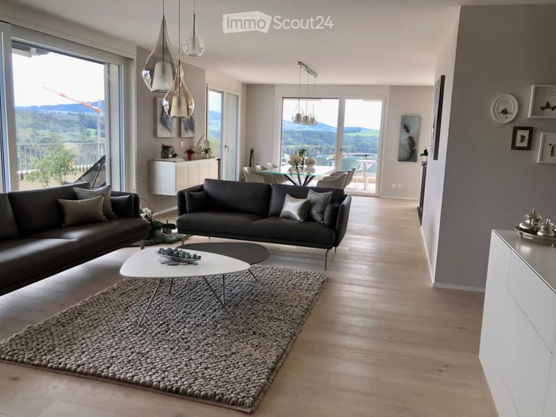 Spacious open-plan living area with large windows offering a scenic view, hardwood flooring, modern gray sofas, a round coffee table with a textured rug, and pendant lighting fixtures.