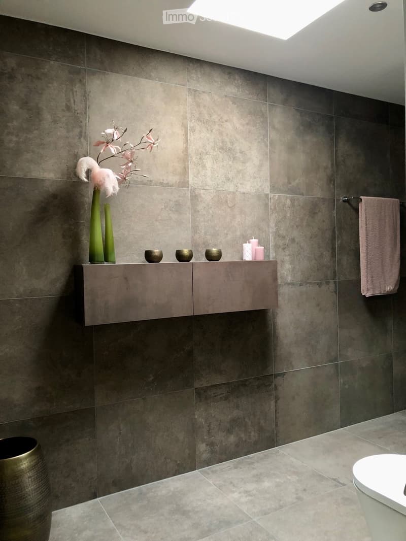 The image shows a modern bathroom with gray tile walls and floors. There is a floating vanity with drawers and decorative items such as vases and a floral arrangement. A pink towel is visible on the side. The room has recessed lighting and appears to be w