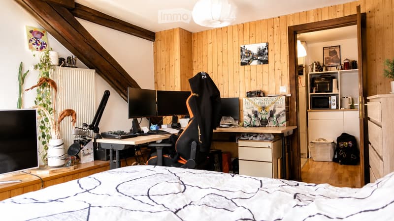 A cozy, multi-functional room with a bed, desk setup, and small kitchen area. The room features wooden walls, a slanted ceiling, and various personal items including a gaming chair, computer monitors, and decorative plants.