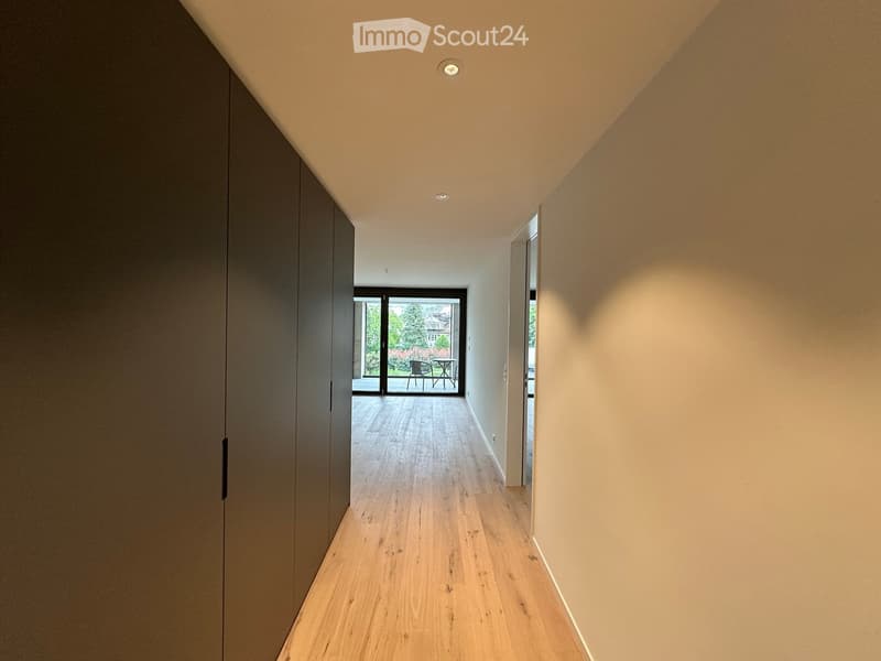 Modern hallway with light wooden flooring, neutral-colored walls, recessed ceiling lights, and a sliding glass door leading to an outdoor area with a small table and chairs.
