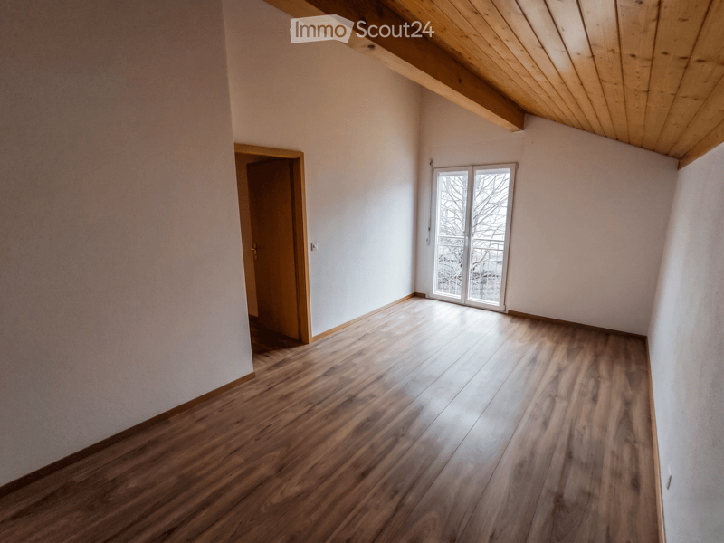 Unfurnished Room. Empty room with wooden flooring, white walls, and a large window with a view of the outside. The room features a wooden door and a slanted wooden ceiling.