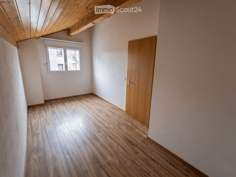 Unfurnished Room. Empty room with wooden flooring, white walls, a wooden door, and a window with white frame. The ceiling has exposed wooden beams.