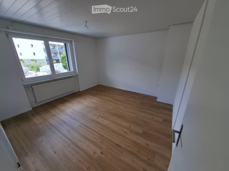 Empty room with wooden flooring, white walls, large window with radiator below, and visible electrical outlets.