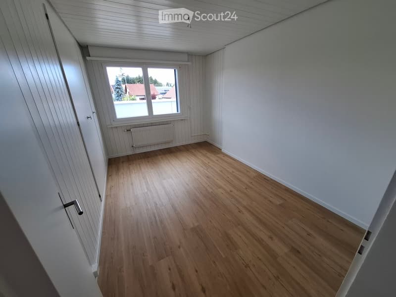 Empty room with wooden flooring, white walls, and a window with a view of the outside. The room features a radiator below the window and a door on the left side.