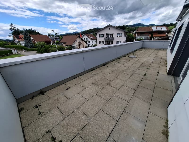 Roof terrace with tiled flooring, low white parapet wall, overlooking a residential neighborhood with houses and trees.