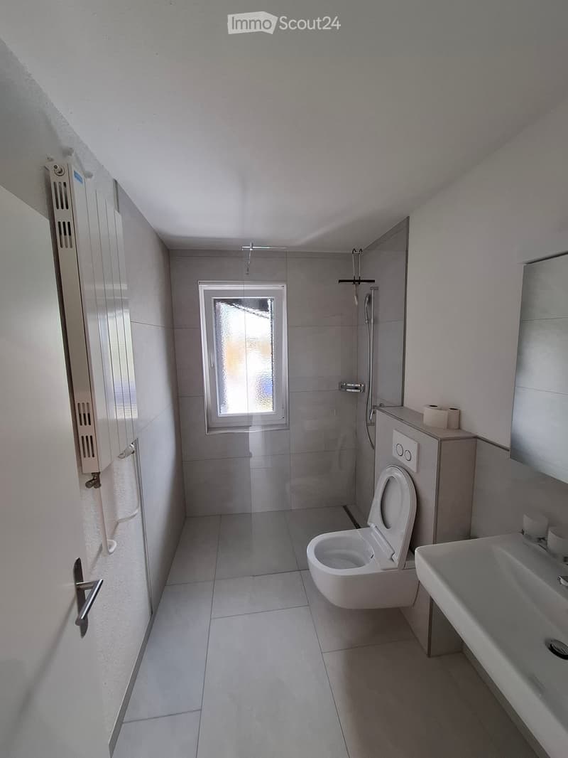 Modern bathroom with white tiles, a toilet, a sink, a shower area, and a frosted window providing natural light.