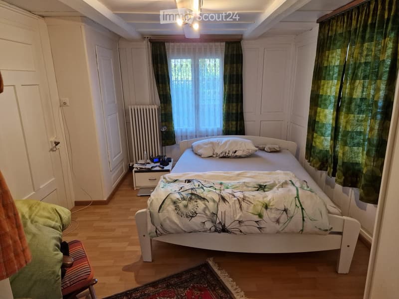 A small bedroom with a single bed, a nightstand, a chair, and a radiator. The room features wooden flooring and a window with green curtains.