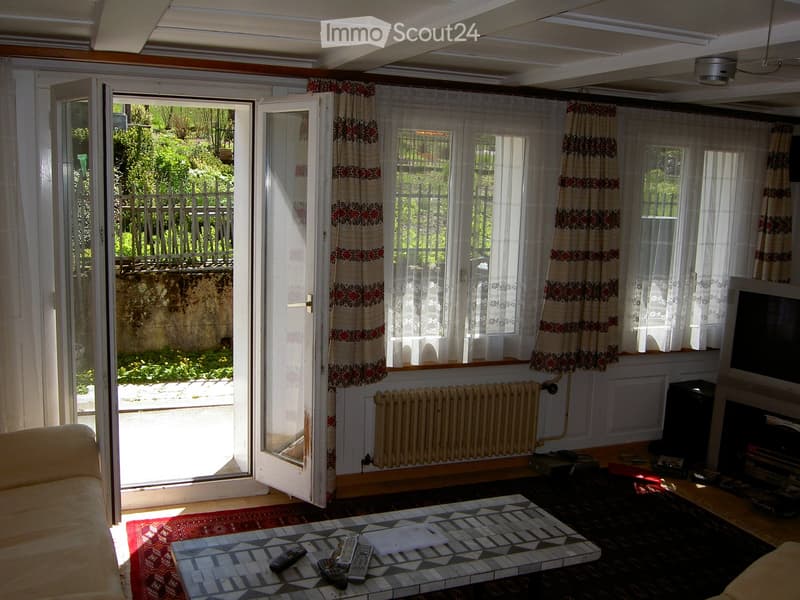 Bright living room with large windows and sliding glass doors leading to a garden, featuring a sofa, coffee table with remote controls, television, and radiator.