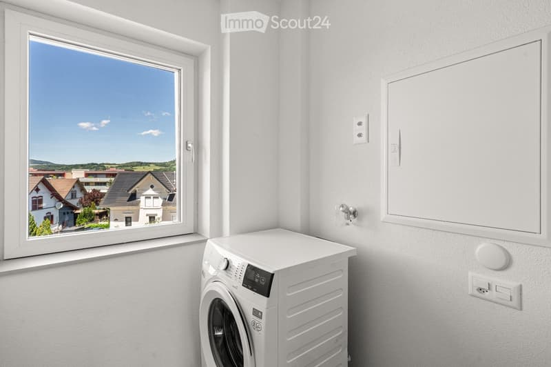 A modern bathroom with a white washing machine, a large window offering a view of the surrounding area, and white walls.