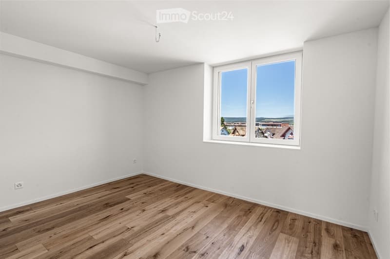 Empty room with white walls, wooden flooring, and a large window offering a view of the outside.