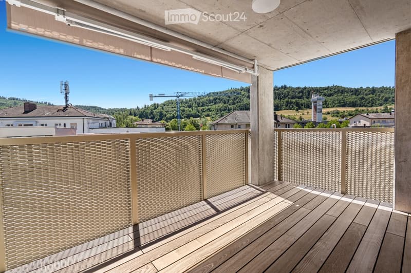Modern balcony with wooden decking, woven metal balustrade, and a view of a green landscape and buildings.