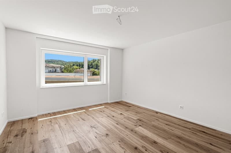 Empty room with light wooden flooring and white walls, featuring a large window with a view of a rooftop and greenery outside.