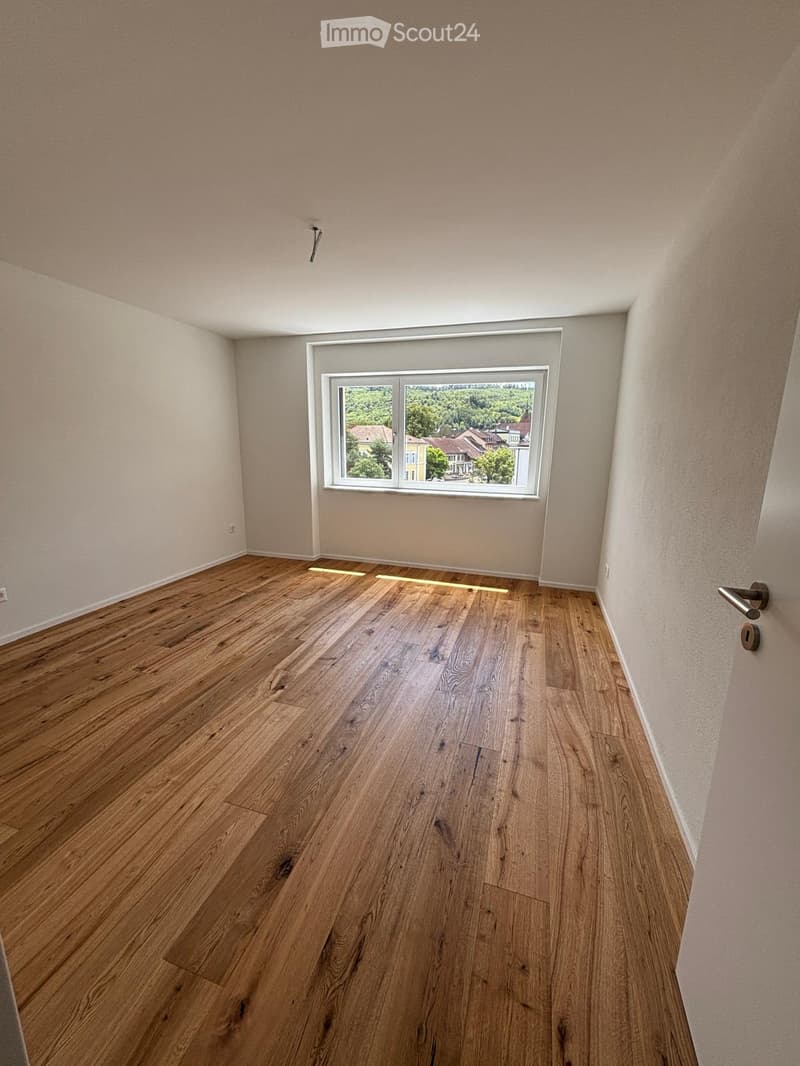 Empty room with wooden flooring and a large window showing a view of trees and houses outside.
