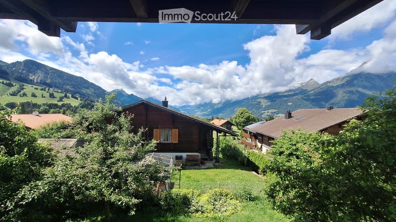 A picturesque view of a wooden chalet surrounded by lush greenery and mountains, with a well-maintained lawn and a bench near the entrance.