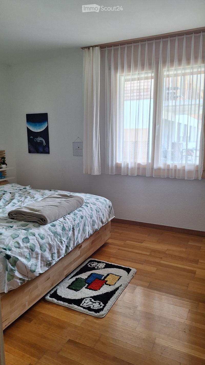 Cozy bedroom with wooden flooring, a bed with a leafy-patterned duvet, a colorful pixelated rug, and a large window with white curtains.