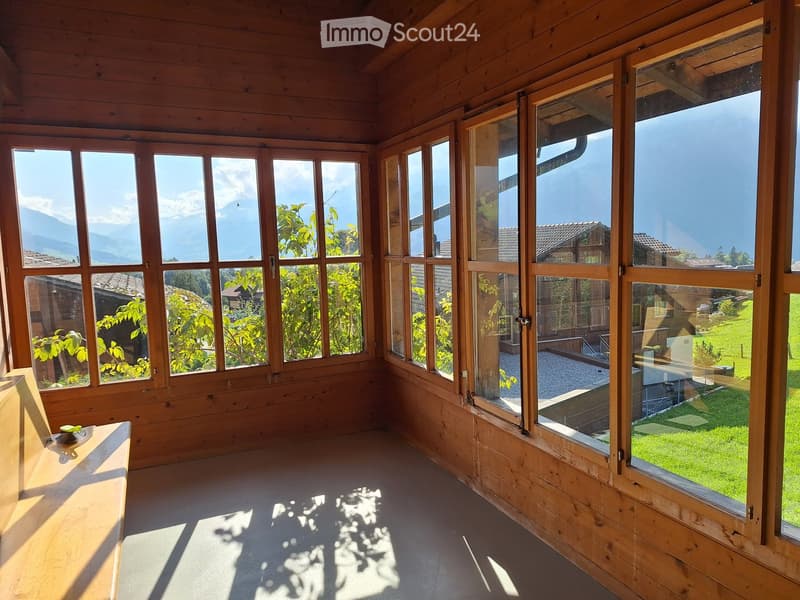 Bright, wooden-framed room with large windows offering views of mountains and greenery. The space is empty, with wooden walls and a smooth floor.