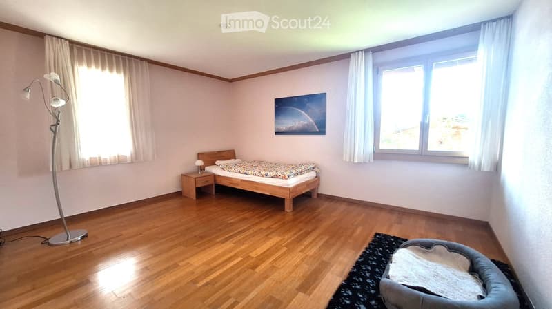 Bright bedroom with wooden flooring, large window with white curtains, bed with colorful bedding, nightstand, floor lamp, and a dog bed in the corner.