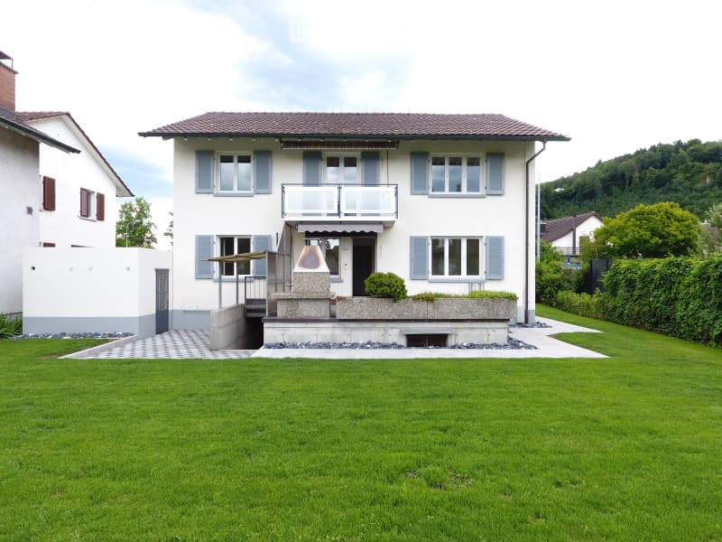 Two-story house with white siding, blue shutters, and a balcony on the second floor. The house has a well-maintained lawn in the front yard and is surrounded by greenery.