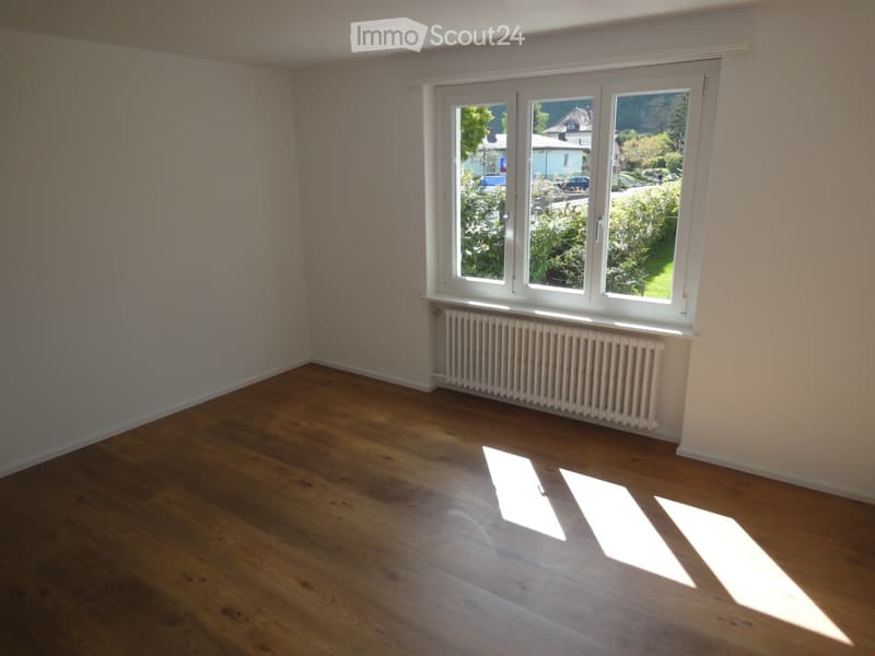 Empty room with wooden flooring, white walls, and large windows showing a view of greenery and houses outside.