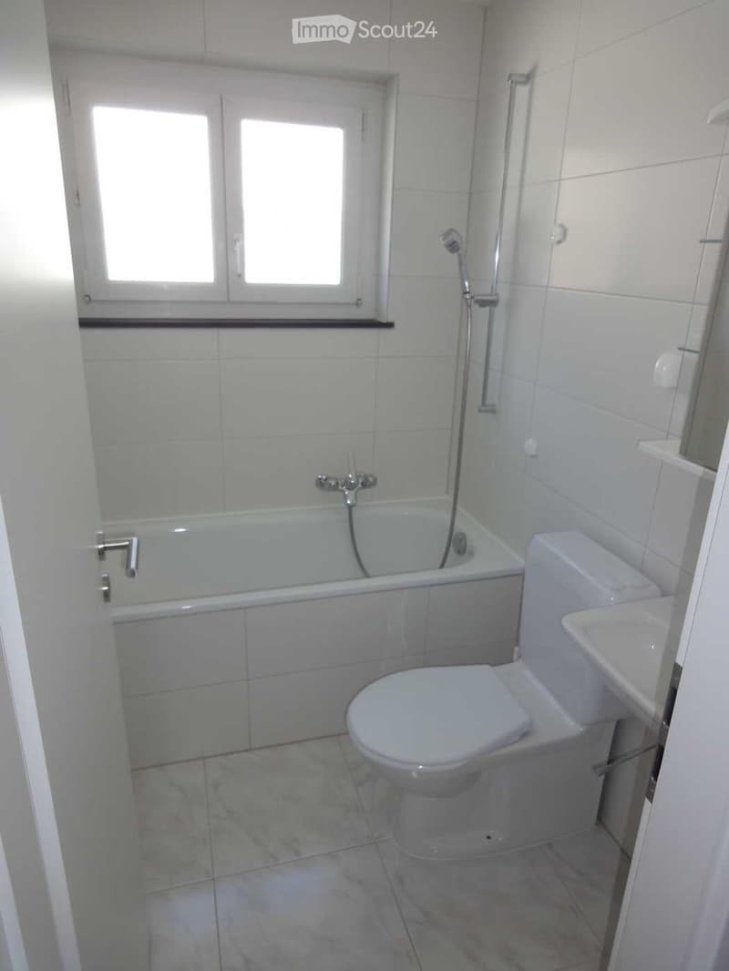 Modern bathroom with white tiles, a bathtub, toilet, sink, and a window above the bathtub.