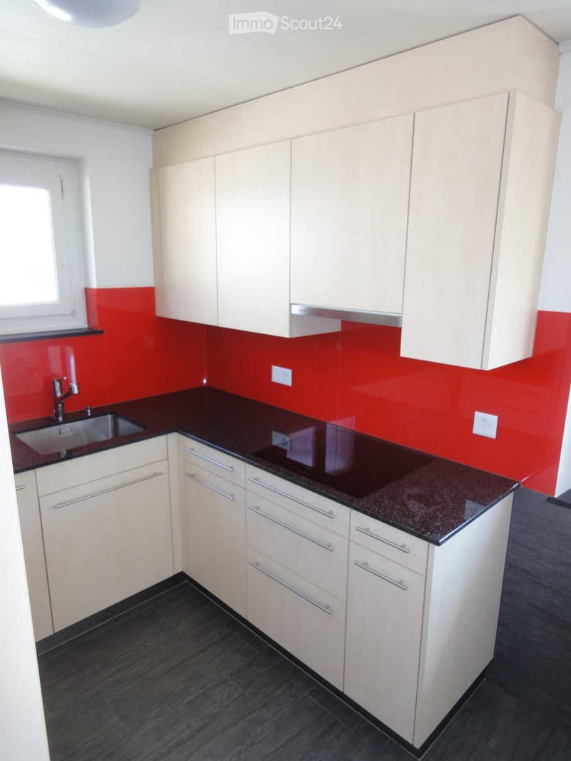Modern kitchen with white cabinets, dark countertops, and a red accent wall. Features a sink with a modern faucet and ample storage space with drawers and cabinets.