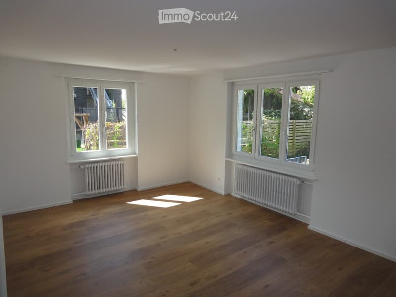 Empty room with wooden flooring, white walls, and large windows offering a view of a garden. Radiators are visible beneath the windows.