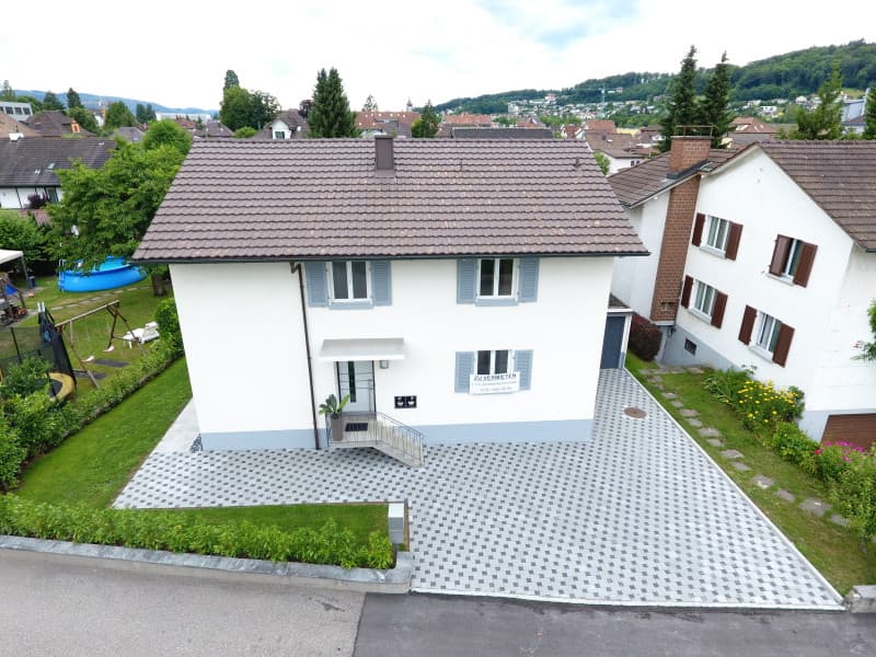 A two-story house with a tiled roof, white exterior, and blue shutters. The house features a small porch with a staircase leading to the entrance, a paved driveway, and a well-maintained garden with a swing set and a small blue pool.