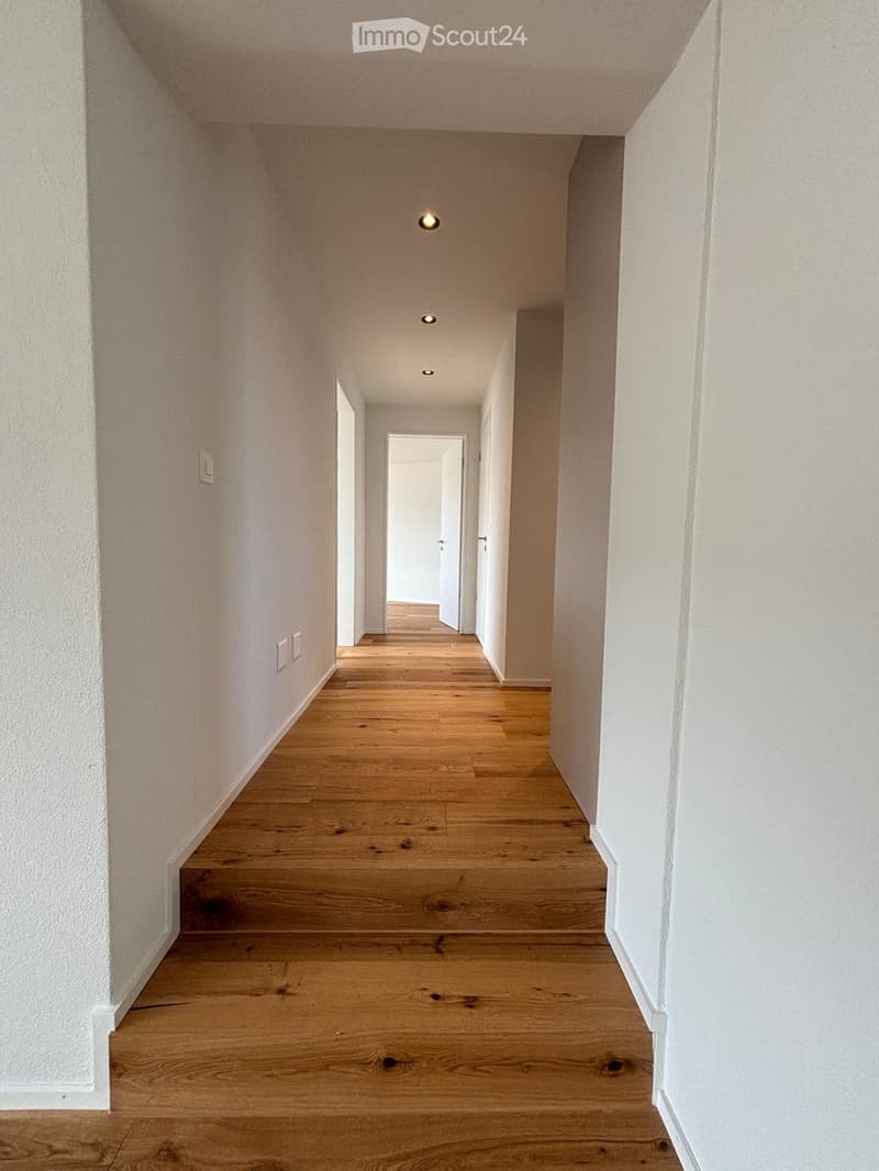 Empty hallway with wooden flooring, white walls, and recessed ceiling lights. Doors visible at the end.