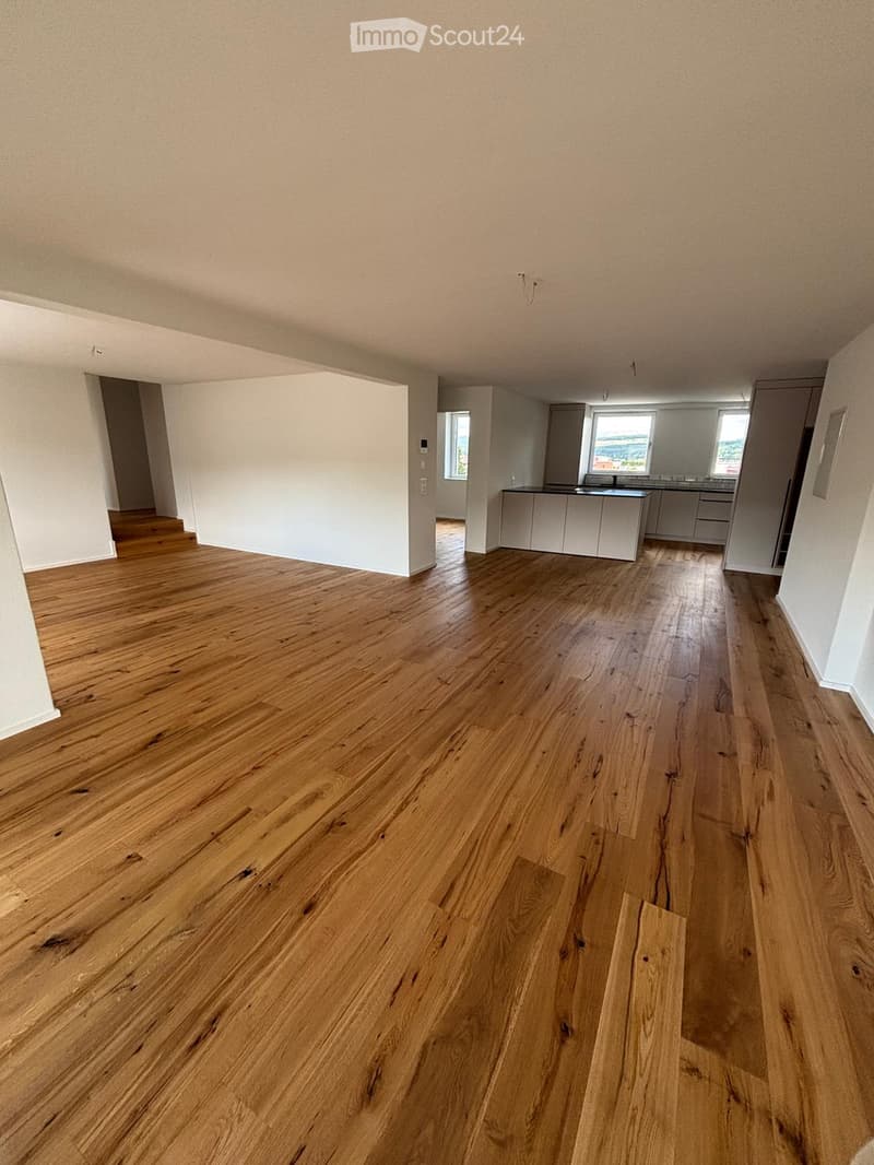 Empty room with wooden flooring and white walls, featuring an open kitchen area with cabinets and countertops visible in the background.