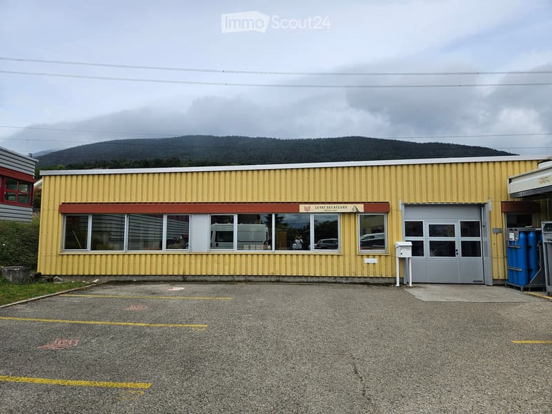 Single-story yellow building with large windows and a grey garage door, located in a parking lot with a mountain view in the background.
